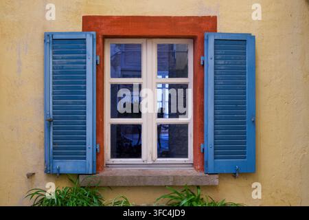 Frontale Nahaufnahme eines charmanten traditionellen Fensters mit blauen Holzläden und einem rot gestrichenen Rahmen auf einer gelben verputzten Wand in Gandria Stockfoto