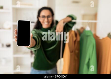 Lächelnde junge Frau hält Smartphone mit leerem Bildschirm in modernem Kleiderschrank, Mockup-Konzept Stockfoto
