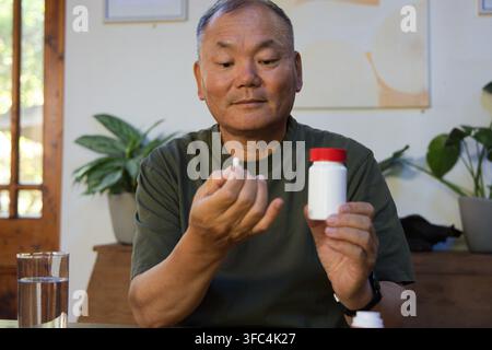 Älterer asiatischer Mann hält eine weiße Pille und eine rotkappen Flasche, bereitet ein Glas Wasser am Esstisch zu Stockfoto