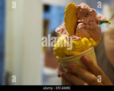 Nahaufnahme einer Hand, die einen Pappbecher mit Löffeln voll Erdbeer- und Mangogelato hält, der mit einem Waffelkeks gekrönt ist. Stockfoto
