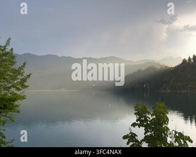 Weicher Abendnebel legt sich über einen ruhigen Bergsee, während die Sonne hinter bewaldeten Hügeln in der Ferne untergeht. Stockfoto