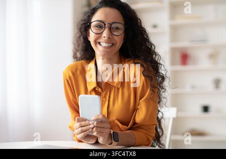 Lächelnde junge Frau hält Smartphone im Büro, glücklicher Ausdruck, Geschäftskonzept Stockfoto
