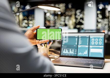 afroamerikanischer Mann, der im modernen Raum mit Green Screen auf dem Handy arbeitet und Online-Einblicke im Strategieraum überprüft. Männlicher General Manager, der sich um die Betriebsverwaltung kümmert. Stockfoto