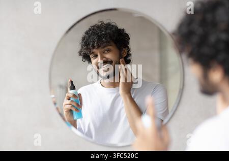 Glücklicher junger Mann, der ein Hautpflegeprodukt vor dem Spiegel zu Hause anwendet, Wellness-Konzept Stockfoto