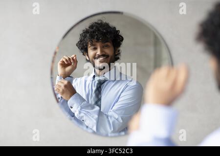 Lächelnder junger südasiatischer Mann, der vor dem Spiegel im Büro seine Uhr anpasst, Geschäftskonzept Stockfoto