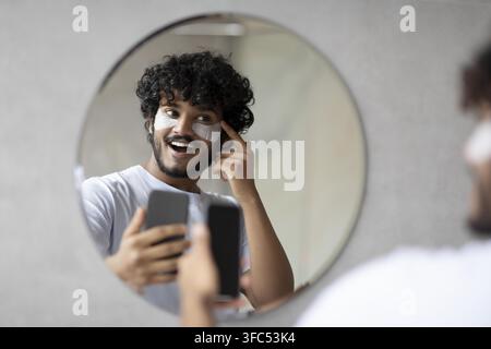 Glücklicher junger Mann, der vor dem Spiegel zu Hause eine Gesichtsmaske aufträgt, Hautpflegeroutine Stockfoto