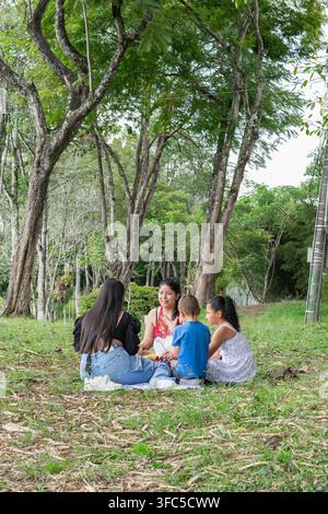Mütter und Kinder genießen ein Picknick, sitzen im Kreis auf dem Gras, umgeben von hohen Bäumen im Park Stockfoto