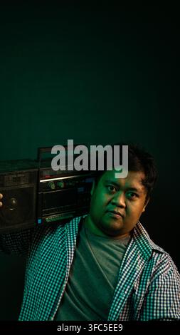 Ein junger asiatischer männlicher Model steht selbstbewusst und hält einen Retro-Boombox auf einer Schulter vor einem dunklen Hintergrund, der von dramatischem grünem und warmem Licht beleuchtet wird Stockfoto