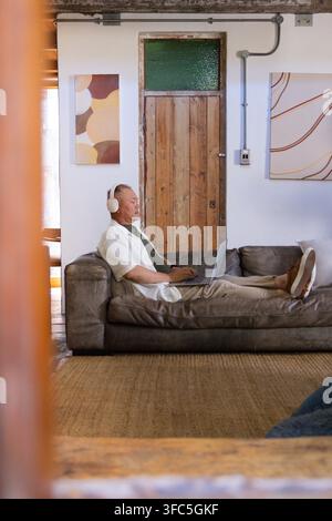 Seniorer asiatischer Mann, der auf einem niedrigen grauen Ledersofa liegt, Kopfhörer trägt und zu Hause einen Laptop benutzt Stockfoto