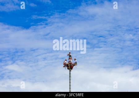 Papantla Flyers führen oben auf dem Ritualpfahl eine Vorführung durch, um sich auf den alten Abstieg vorzubereiten, vor einem wolkenverhangenen blauen Himmel. Stockfoto