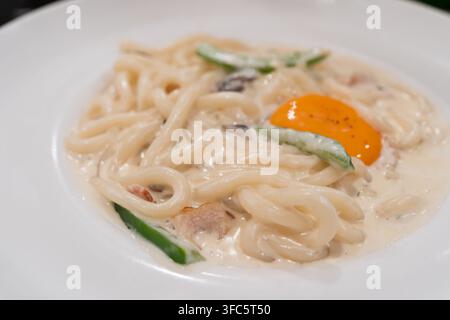 Kremige Carbonara-Udon-Nudeln serviert in einer reichhaltigen weißen Sauce mit Pilzen, Speck und frischen Gurkenscheiben, belegt mit einem weichen orangefarbenen Eigelb. Stockfoto