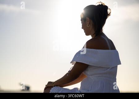 Eine Frau in einem weißen Kleid steht in der Sonne mit dem Rücken zur Kamera. Sie blickt zum Horizont hinaus. Ihr Haar weht im Wind Stockfoto
