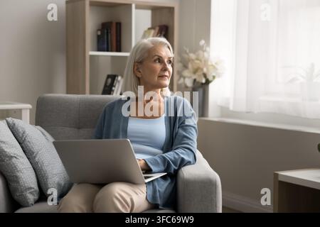 Ältere Frau, die von zu Hause aus arbeitet, sitzt in einem Sessel mit Laptop Stockfoto
