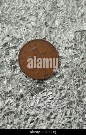 Britische Zwei-Pence-Münze, die auf zerknittertem Aluminiumfolie steht, kontrastiert Kupfermünzen mit einem reflektierenden silbernen Metallhintergrund Stockfoto