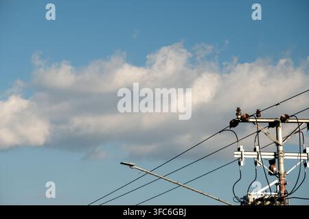 Versorgungsinfrastruktur, insbesondere Strommasten mit Transformatoren und Stromnetzen vor einem bewölkten blauen Himmel. Stockfoto