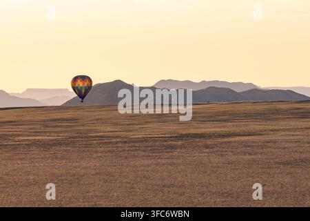Heißluftballon schwebt sanft über die weite Wüstenlandschaft von Sossusvlei bei Sonnenaufgang und zeigt die weiten Ebenen und fernen Berge unter einem Stockfoto