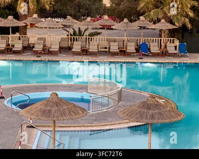 Resort-Schwimmbad mit klarem blauem Wasser und leeren Sonnenliegen unter Strohschirmen, die Entspannung und Urlaub versprechen. Ammoudara Beach, Heraklion Stockfoto