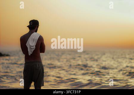 Ein einsamer Mann erscheint als Silhouette, während er das warme Sonnenlicht über dem Horizont beobachtet. Stockfoto