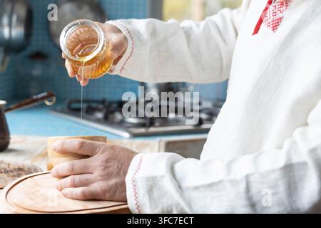 Ethnisches Leinenhemd am Mann. Ein bärtiger Mann in der Küche gießt Honig in eine Tasse. Dessert zubereiten in einer rustikalen Küche. Stockfoto
