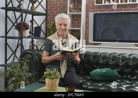 Ein älterer Mann sitzt auf einem dunkelgrünen Ledersofa in einem gemütlichen Wohnzimmer und liest ein aufgeschlagenes Buch. Der Raum verfügt über eine Ziegelwand, Regale und eine Stockfoto