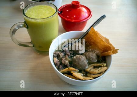 Grünes Smoothy-Getränk neben einer Schüssel mit Fleischbällchen. Gebratene Snacks und ein Löffel ruhen in der Schüssel. Ein Behälter mit rotem Deckel steht im Hintergrund. Simplizistisches Holz Stockfoto