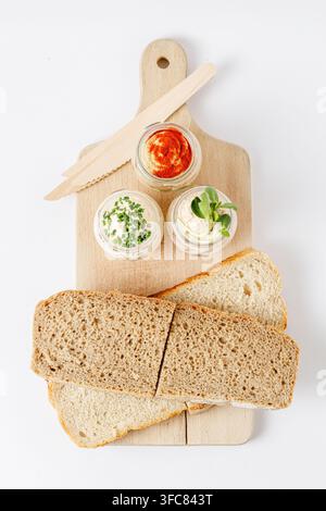 Brot und verschiedene Aufstriche in Gläsern auf Schneidebrett isoliert Stockfoto
