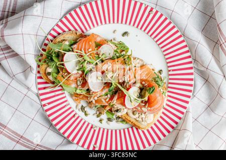 Gourmet-Lachs-Toast mit Frischkäse, Rettich, Rucola und Samen auf einem rot gestreiften Teller Stockfoto
