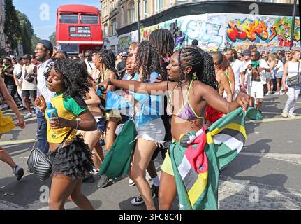 Karibische Karnevalsbesucher mit einer Flagge von Saint Kitts und Nevis passieren einen roten Londoner Bus während der Hauptparade des Notting Hill Karnevals 2025 Stockfoto