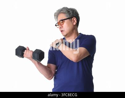Älterer koreanischer Mann, der beim Training mit einem Hantel gegen einen weißen Hintergrund Schulterschmerzen verspürt. Stockfoto