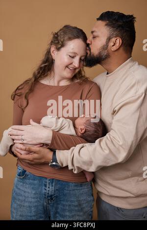 Interrassisches Familienporträt mit liebevollen Eltern, die ein Neugeborenes halten, Vater küsst die Stirn der Mutter Stockfoto