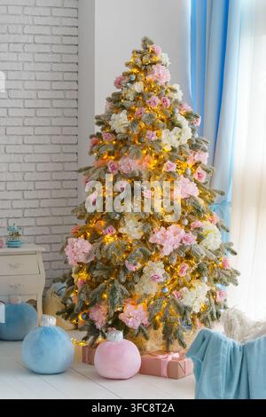 Festlicher Weihnachtsbaum dekoriert mit rosa und weißen Blumen, silbernen Ornamenten und warmen Lichtern in einem hellen, gemütlichen Interieur mit pastellfarbenen Geschenken. Festliche Ästhetik Stockfoto