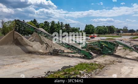 Schwere Maschinen arbeiten auf einer Baustelle, um Kies zu verarbeiten und Haufen zu schaffen. Die Ausrüstung fügt sich in die natürliche Landschaft unter einem blauen Himmel ein mit Stockfoto