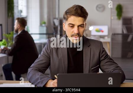 Ernsthafter Geschäftsmann im Anzug, der an einem modernen offenen Büroraum an einem Laptop arbeitet Stockfoto