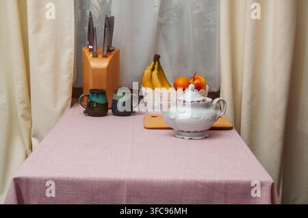 Eine charmante Küchenszene mit einer Sammlung von Keramiktassen, einem dekorativen Teekessel, frischem Obst und einem Messerblock, die auf einem Tisch arrangiert sind, der mit abgedeckt ist Stockfoto