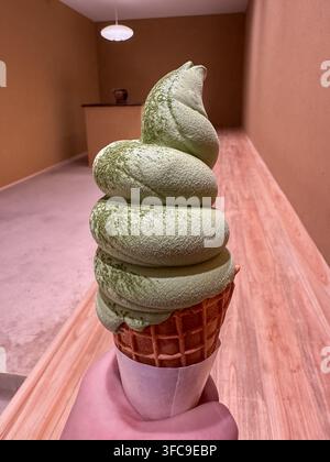 Matcha-Grüntee-Softeis in einer Waffeltasche, die von einer Hand gehalten wird, vor einem minimalistischen rosa Interieur. Stockfoto