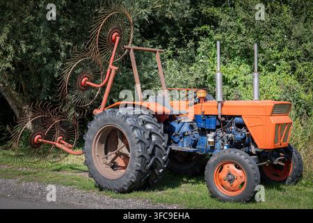 Ein alter orangefarbener Traktor mit einem großen, rostigen Heurake ist auf einem grasbewachsenen Feld mit Bäumen im Hintergrund geparkt Stockfoto