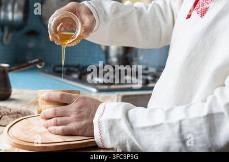 Ethnisches Leinenhemd am Mann. Ein bärtiger Mann in der Küche gießt Honig in eine Tasse. Dessert zubereiten in einer rustikalen Küche. Stockfoto