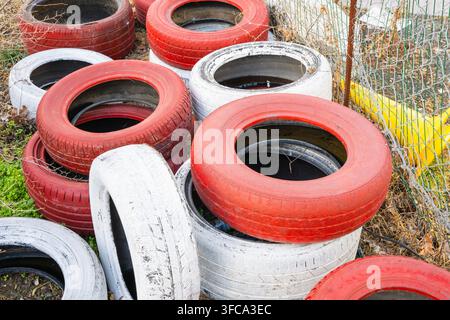 Ein verstreuter Haufen alter Autoreifen, rot und weiß bemalt, liegt im Freien auf überwuchertem Gras neben einem Drahtgitterzaun Stockfoto