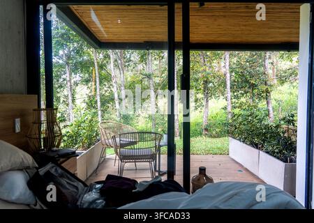 Das helle Schlafzimmer öffnet sich zu einer hölzernen Terrasse mit Glastüren, Korbsesseln und üppigem Grün direkt vor dem Hotel. Weiche Bettwäsche, Tageslicht und ein sauberer, moderner Look Stockfoto
