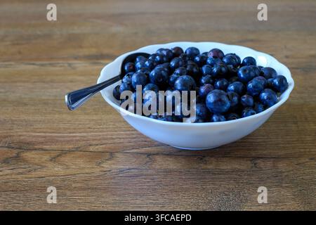 Weißer Schüssel, der mit saftigen Blaubeeren überquillt, und ein Teelöffel, der auf einer strukturierten Holzoberfläche platziert ist. Konzept von gesunder Ernährung, frischen landwirtschaftlichen Produkten und sei Stockfoto