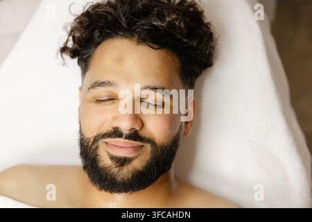 Bärtiger Mann mit geschlossenen Augen, der lächelt, während er eine entspannende Spa-Gesichtsbehandlung genießt, liegt auf einem weißen Bett in weichem natürlichem Licht Stockfoto