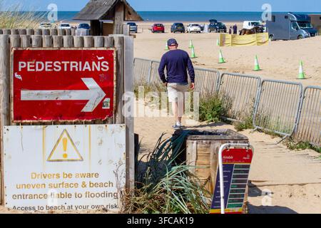 Ein Fußgänger-Direktionsschild und Warnhinweise markieren einen Strand-eingang, während ein Mann den Weg entlang zu einem sandigen Strandparkplatz geht. Southport, UK. Stockfoto