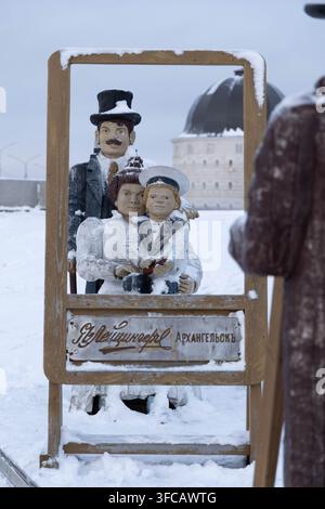 Holzfiguren einer Familie, die in einem Rahmen eines alten Fotostudios posieren, mit der Gostiny-Dvor-Kuppel im Hintergrund im winterlichen Arkhangelsk, Russland. Stockfoto