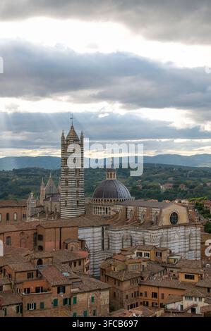 Vertikale Ansicht der Kathedrale von Siena, des gestreiften Glockenturms und der Kuppel, die sich über historischen Dächern erheben, mit Sonnenstrahlen, die dunkle Wolken in Siena, Toskana, Ita durchbohren Stockfoto