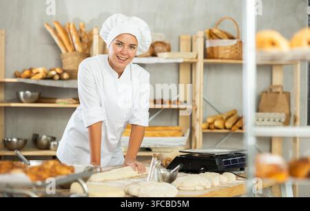 Mädchen arbeitet als Bäckerin in einer Bäckerei, stellt Backwaren aus Teig her. Die Arbeiterin hat aus Zutaten einen Teigklumpen geschaffen und bereitet ihn zum Schneiden vor. Sie steht Stockfoto