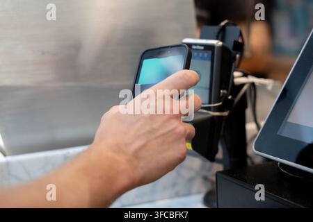 Zuschnittbild einer Männerhand, die ein Smartphone hält, um eine digitale Zahlung mit einer mobilen Geldbörse über kontaktloses Bezahlen an einem POS-Terminal vorzunehmen Stockfoto
