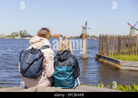 Eine Mutter und ihre Tochter sitzen am Wasser in den Niederlanden, blicken auf Windmühlen und genießen ihren Familientrip an einem sonnigen Tag. Stockfoto