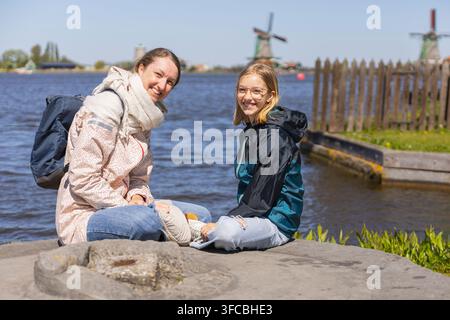 Eine Mutter und ihre Tochter sitzen zusammen am Wasser, genießen ihre Reise in den Niederlanden und blicken auf Windmühlen in der Ferne. Stockfoto