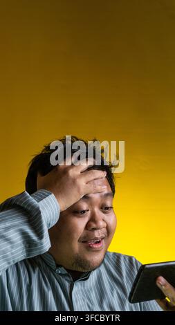 Ein erleichterter junger asiatischer Mann legt eine Hand an die Stirn, während er nach unten auf ein Smartphone blickt, vor einem lebhaften gelben Hintergrund, der ein Gefühl vermittelt Stockfoto