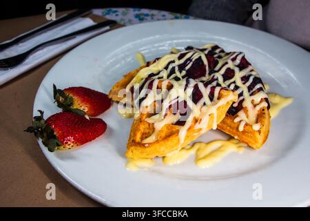Nahaufnahme von goldenen Waffeln, die mit cremiger weißer Sauce und dunklem Beerenkompott überzogen sind, serviert mit frischen Erdbeeren auf einem weißen Teller. Stockfoto